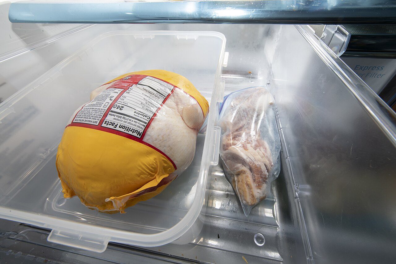 Frango inteiro em embalagem fechada descongelando numa bandeja com tampa na gaveta da geladeira, ao lado de um saco de bacon