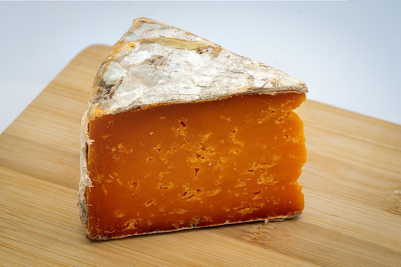 Fatia triangular de queijo cheddar maturado alaranjado com casca, sobre tábua de madeira