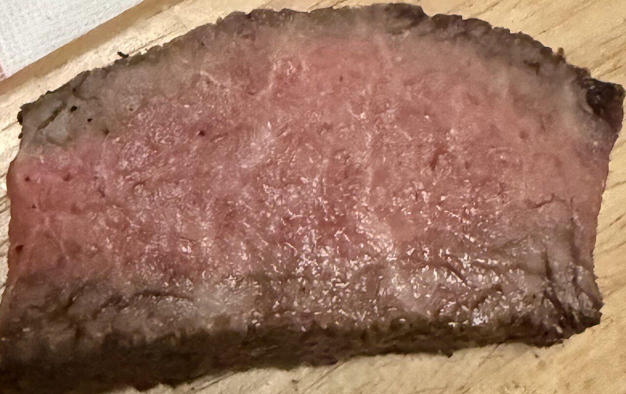Fatia de bife cortada, com o centro rosado, a borda externa marrom escura e as laterais cinzentas, sobre uma tábua de madeira