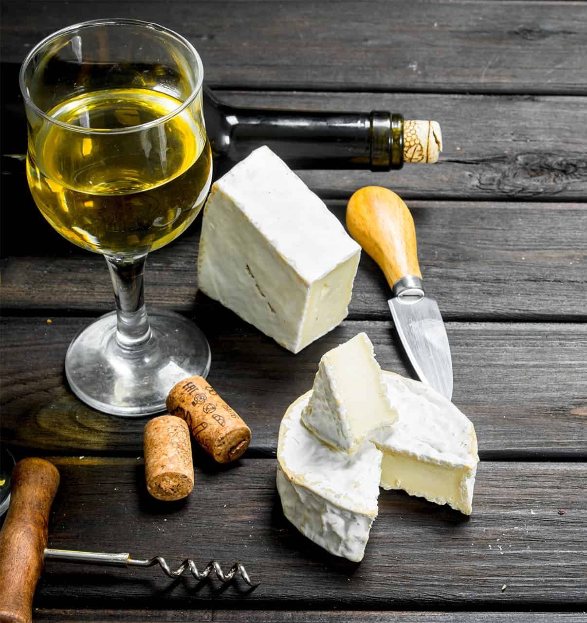Queijo brie em pedaços ao lado de taça de vinho branco, rolhas e saca-rolhas