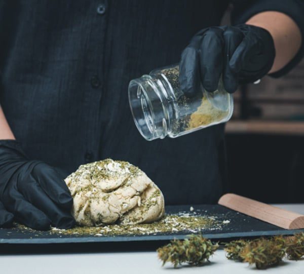 Mãos de luvas pretas polvilham erva moída sobre massa, com flores de cannabis na bancada