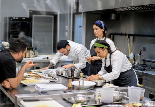 Chef observa cozinheiros de dólmã montando pratos juntos numa cozinha profissional