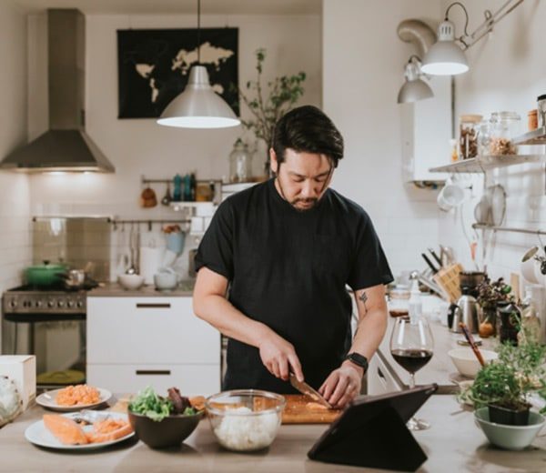 Cozinheiro corta salmão numa cozinha doméstica, com tablet e taça de vinho tinto na bancada