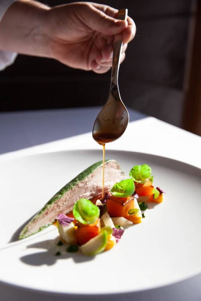 Chef regando com colher o molho sobre peixe e legumes num prato branco