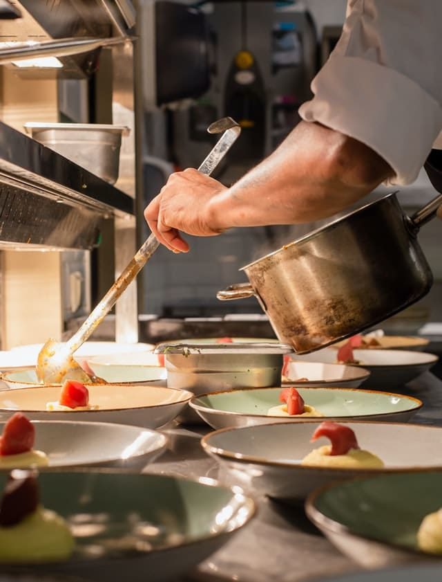 Cozinheiro servindo purê com concha em pratos enfileirados numa cozinha profissional