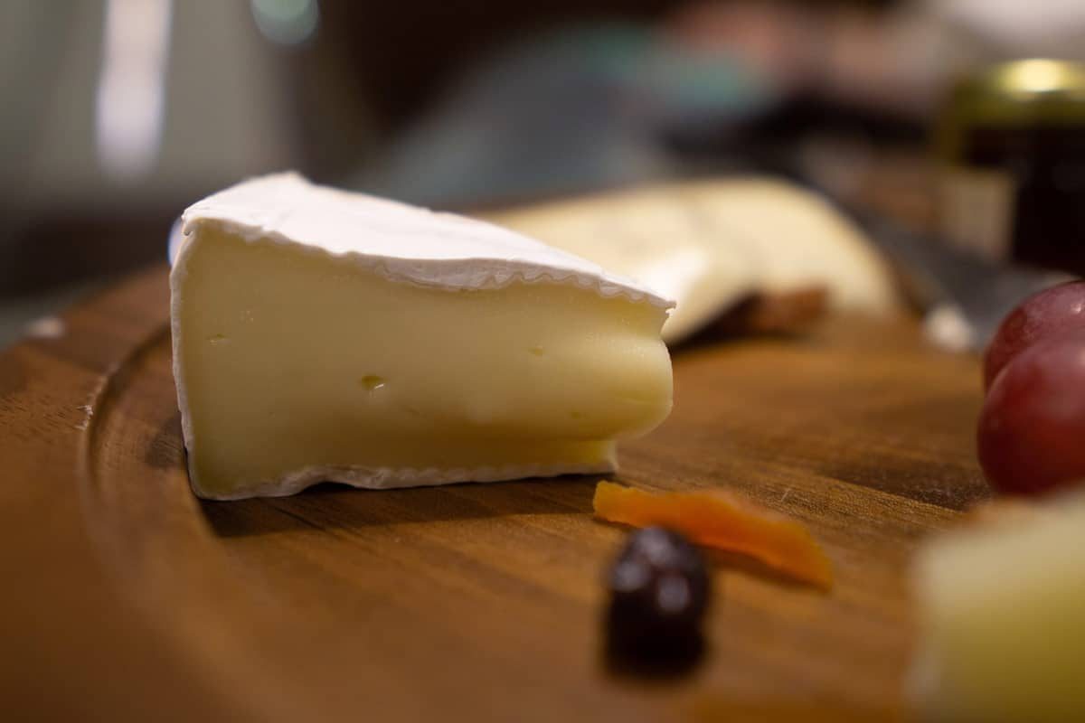 Fatia de queijo brie de casca branca sobre tábua de madeira com uvas e frutas secas