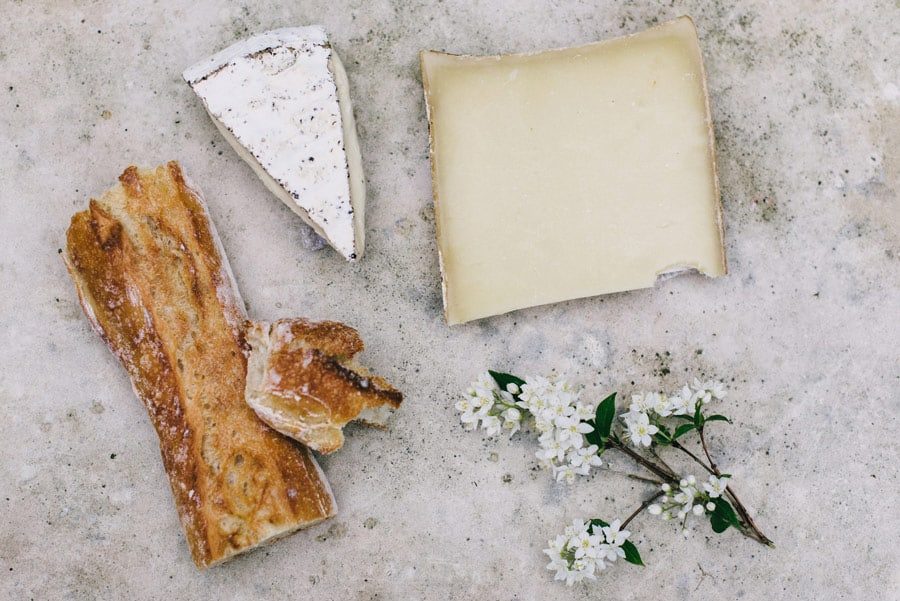 Fatia de queijo brie e pedaço de queijo curado ao lado de baguete e flores brancas