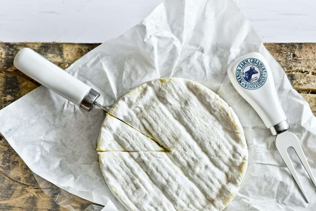Queijo brie inteiro com uma fatia cortada sobre papel manteiga, com faca e garfo de queijo